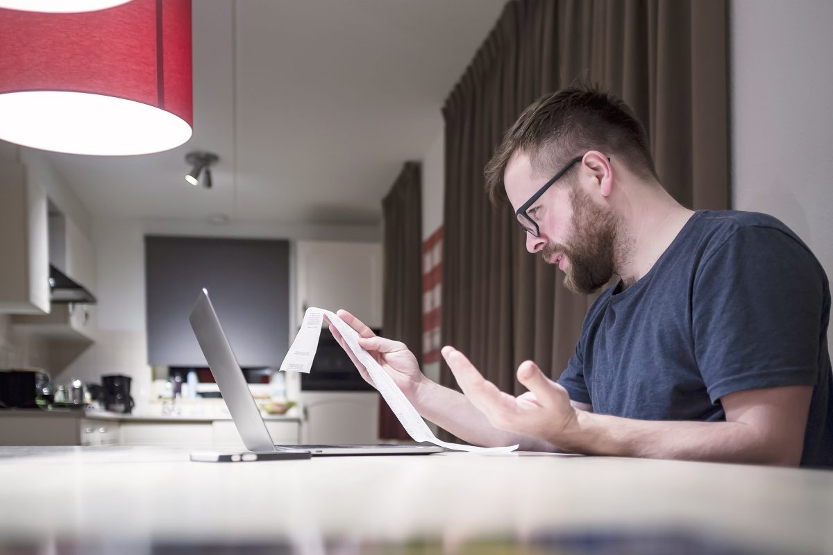 man managing finances sitting near laptop looking receipt being surprise unhappy
